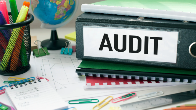 Ilustratīvs attēls par auditu – uz darba galda redzamas dokumentu mapes, piezīmju bloki un biroja piederumi; uz vienas mapes izvietots uzraksts “AUDIT”.