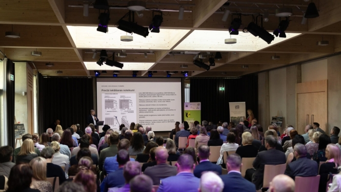 Konferences dalībnieki sēž zālē un klausās prezentāciju. Uz skatuves runātājs prezentē slaidu par precīzi ielāšanas noteikumiem; zāle ir piepildīta ar apmeklētājiem.