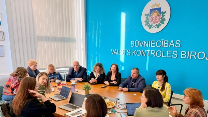 Darba sanāksme Būvniecības valsts kontroles birojā. Ap galdu sēž vairāki dalībnieki, kuri pārrunā darba jautājumus; uz galda redzami portatīvie datori, dokumenti un pierakstu materiāli. Fonā – BVKB nosaukums un Latvijas valsts ģerbonis.