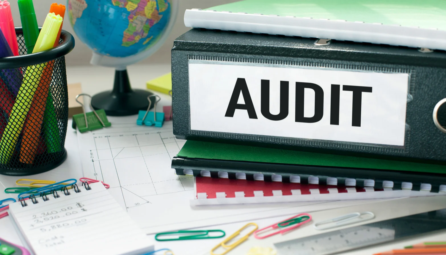 Ilustratīvs attēls par auditu – uz darba galda redzamas dokumentu mapes, piezīmju bloki un biroja piederumi; uz vienas mapes izvietots uzraksts “AUDIT”.