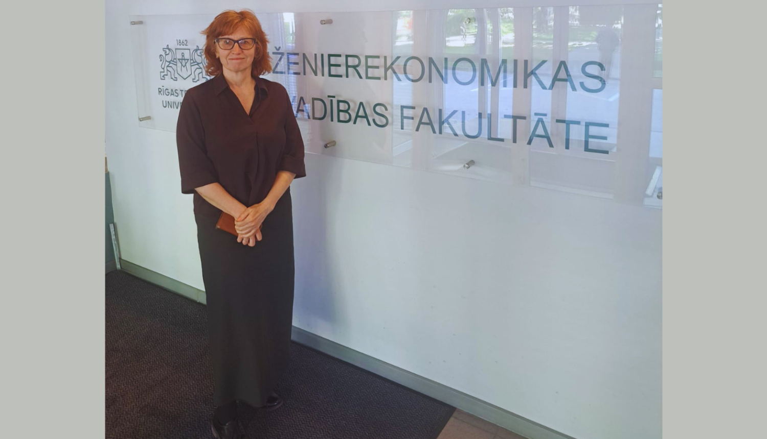 Fotogrāfijā redzama Baiba Vītoliņa, Būvniecības valsts kontroles biroja direktore. Viņa stāv ar saliktām rokām un klusu smaidu Rīgas Tehniskās universitātes telpās, blakus stikla sienai ar uzrakstu latviešu valodā "INŽENIEREKONOMIKAS UN VADĪBAS FAKULTĀTE".