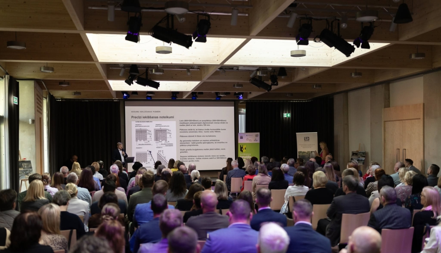 Konferences dalībnieki sēž zālē un klausās prezentāciju. Uz skatuves runātājs prezentē slaidu par precīzi ielāšanas noteikumiem; zāle ir piepildīta ar apmeklētājiem.