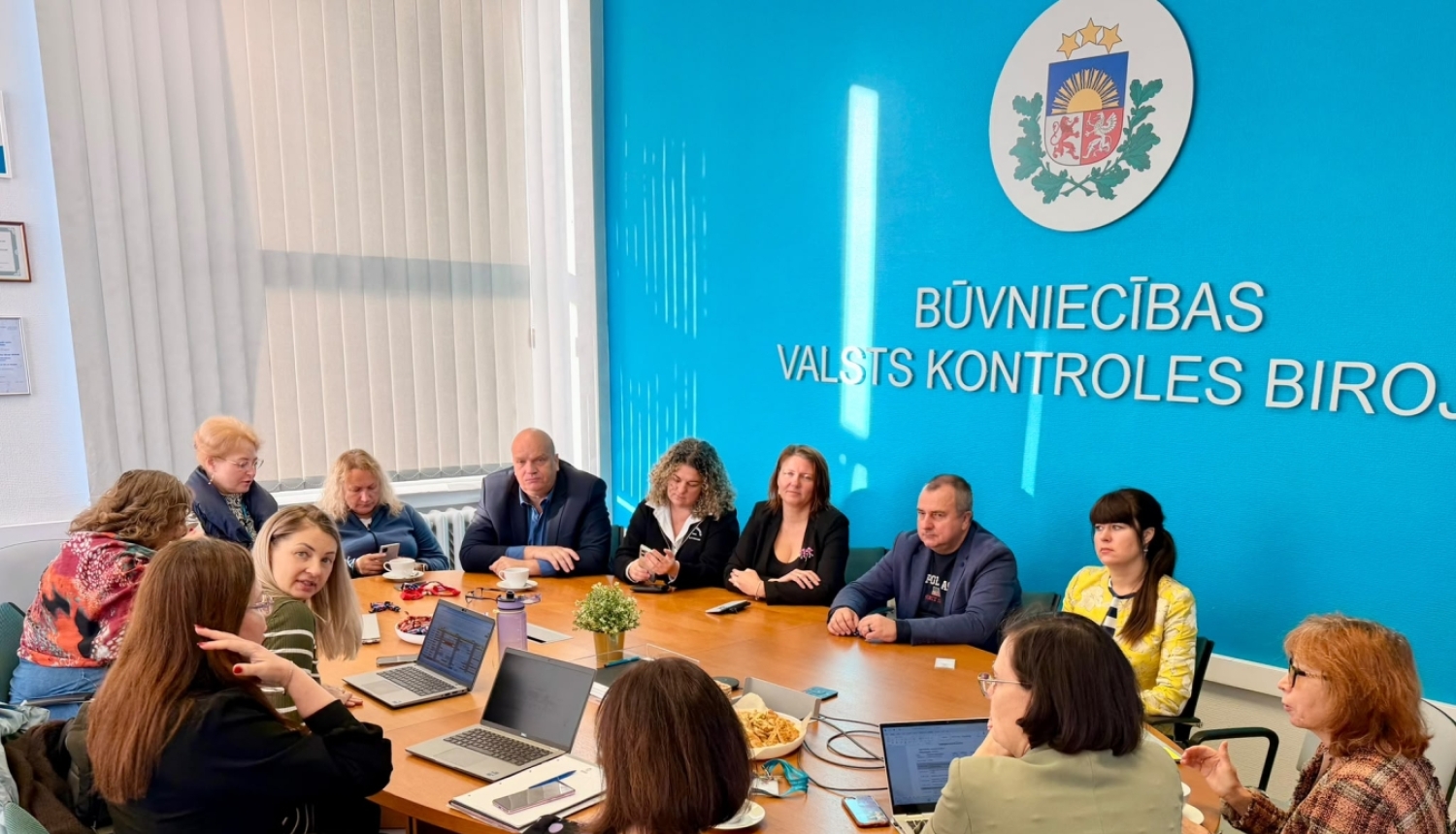 Darba sanāksme Būvniecības valsts kontroles birojā. Ap galdu sēž vairāki dalībnieki, kuri pārrunā darba jautājumus; uz galda redzami portatīvie datori, dokumenti un pierakstu materiāli. Fonā – BVKB nosaukums un Latvijas valsts ģerbonis.