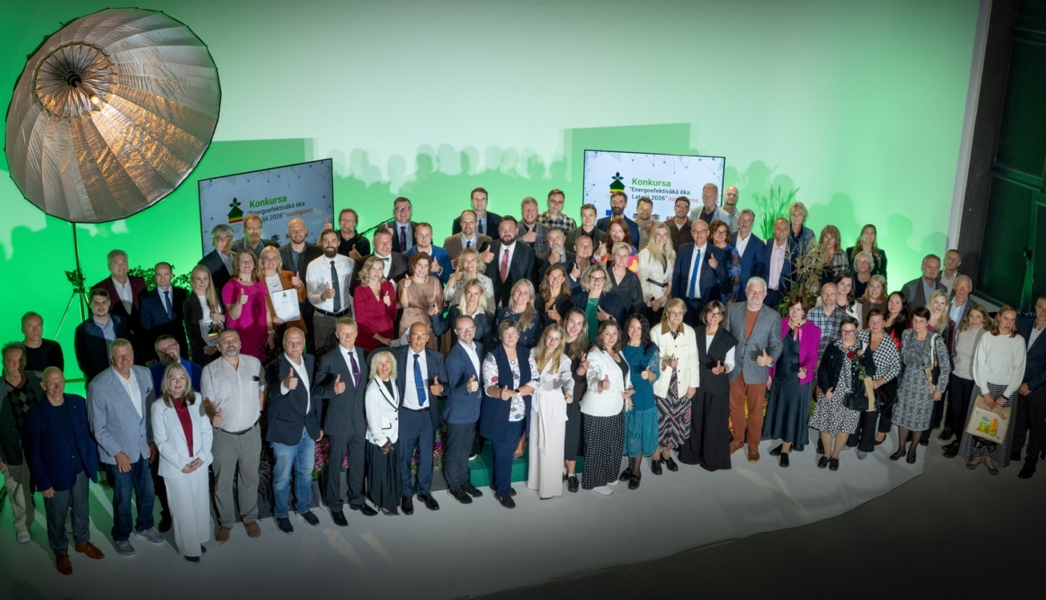 Kopbilde no konkursa “Energoefektīvākā ēka Latvijā 2026” apbalvošanas pasākuma. Uz skatuves kopā fotografējas konkursa dalībnieki, laureāti un organizatoru pārstāvji; fonā redzami konkursa vizuālie materiāli.